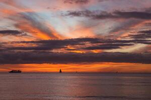 Vibrant sunset with orange, pink, and purple hues over calm ocean waters. A cruise ship and sailboats, including one with a triangular sail, are visible. photo