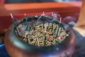 Close up of a bronze incense burner filled with ash and glowing incense sticks, emitting smoke. Vibrant red and orange hues suggest a temple setting. photo
