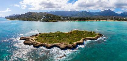 Aerial view of a small, rocky island with greenery and walking paths, surrounded by turquoise waters near Oahu's coastline with beaches and mountains. photo
