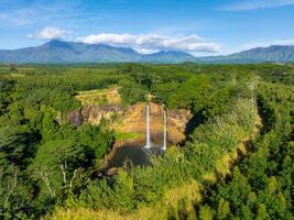 Twin waterfalls cascade into a tranquil pool, surrounded by lush vegetation and dense forests, with mountains visible under a clear blue sky. photo