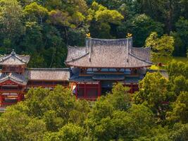 The Byodo In Temple in Oahu, Hawaii, features red wooden structures, a curved tiled roof, and golden accents, set amidst dense trees and greenery. photo