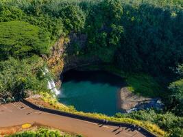 Wailua Falls cascades into a circular pool, surrounded by steep cliffs and vibrant greenery. A paved road and visitors are visible in the foreground. photo