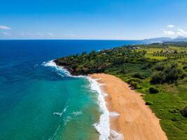 Aerial view of a curved golden sand beach on Kauai Island, Hawaii, bordered by turquoise waters, lush green vegetation, and scattered distant structures. photo
