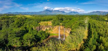 Twin waterfalls of Wailua Falls cascade into a serene pool, surrounded by dense forest and mountainous terrain under a bright blue sky with clouds. photo