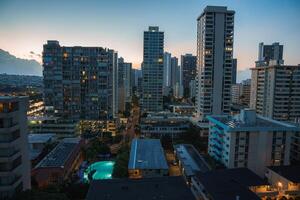 View of Honolulu at dusk featuring illuminated high rise buildings, a swimming pool in the foreground, and distant mountains on the left. photo