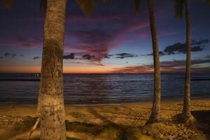 A tranquil beach scene at sunset with vibrant orange, pink, and purple skies. Palm trees frame the view, and small boats float on the calm ocean. photo