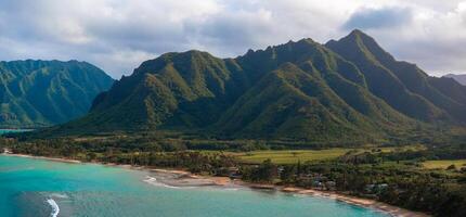 Aerial view of Oahu, Hawaii, showing green mountains with sharp ridges, a turquoise shoreline, sandy beach, small settlement, and dense vegetation. photo