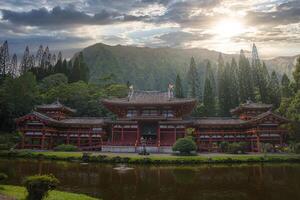 The Byodo In Temple stands amidst lush greenery, a reflective pond, and the Ko olau mountain range under a partly cloudy sky in Oahu, Hawaii. photo
