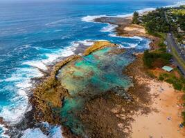 Aerial image of a turquoise lagoon with rugged rocks, a sandy beach with visitors, and a road lined with greenery on Oahu island, Hawaii. photo