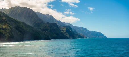 Towering green cliffs of the Napali Coast meet the vibrant blue ocean under a bright sky with scattered clouds, illuminated by soft sunlight. photo