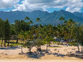Golden sandy beach on Oahu island with palm trees, lush vegetation, and the Ko olau mountain range under a partly cloudy sky in the background. photo