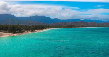 Aerial view of a sandy beach with turquoise waters on Oahu island, Hawaii, bordered by trees and backed by lush green and brown mountains under a cloudy sky. photo