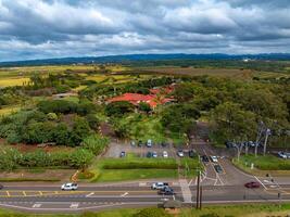 Aerial image of Dole Plantation on Oahu Island, Hawaii, with red roofed buildings, tropical trees, farmland, rolling hills, and a cloudy sky. photo