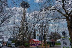 The Space Needle stands tall above leafless trees, with a red roofed kiosk, signs, and parked vehicles in an urban park setting under a cloudy sky. photo