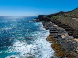 Rugged cliffs and rocky shores meet vibrant blue ocean waves in Oahu, Hawaii. A winding road runs along the hillside under a clear sky. photo