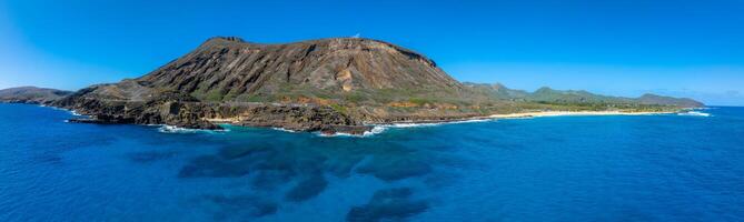 Aerial view of a volcanic mountain with rugged slopes near the shoreline on Oahu Island, Hawaii, featuring sandy beaches, a winding road, and clear skies. photo