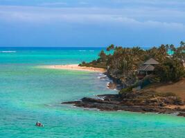 Turquoise waters meet a sandy beach lined with palm trees in Oahu, Hawaii. A house sits near rocky terrain, with people visible enjoying the beach. photo