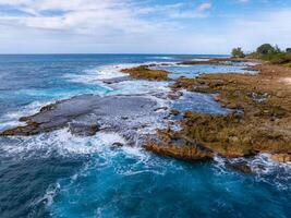 Rugged rocky shoreline of Oahu, Hawaii, with tide pools, turquoise waters, sparse greenery, and a partly cloudy sky in the background. photo