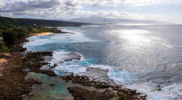 Aerial view of Oahu's rocky coastline with turquoise tide pools, lush greenery, distant mountains, and sunlight reflecting on the ocean under a cloudy sky. photo