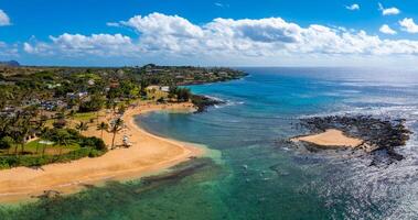 Golden sandy beach on Kauai island, Hawaii, bordered by greenery and palm trees, with turquoise waters, visitors, and a residential area in the background. photo