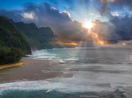 Aerial view of Kauai Island showing Napali Coast cliffs, lush greenery, sandy beach, turquoise waves, and sunlight breaking through clouds. photo