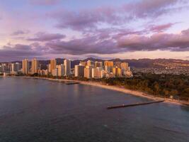 Aerial view of Waikiki Beach in Honolulu, Oahu, Hawaii, showing high rise buildings, calm ocean waters, sandy beach, and a mountainous backdrop at sunset. photo