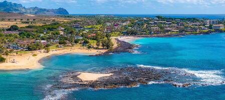 Aerial view of Kauai island's coastline featuring turquoise waters, sandy beaches, palm trees, a small town, and distant hills in the background. photo