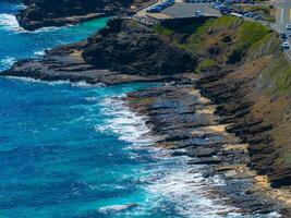 Rugged black volcanic cliffs meet turquoise Pacific waters in Oahu, Hawaii. A parking area and winding road sit atop the cliffs, bordered by greenery. photo
