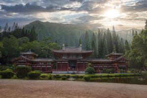 The Byodo In Temple in Oahu, Hawaii, surrounded by greenery, tall pines, and mountain ridges, with a tranquil pond reflecting the serene scene. photo