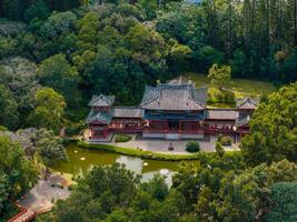 Aerial view of the Byodo In Temple in Oahu, Hawaii, featuring traditional Japanese architecture, a reflective pond, and dense forest surroundings. photo