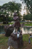 A bronze statue of a young girl stands on a rock near a pond, surrounded by pigeons, with lush trees and grass in a tranquil park setting in Hawaii. photo