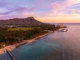 Aerial view of Waikiki Beach in Oahu, Hawaii, at sunset with Diamond Head Crater in the background, turquoise waters, sandy shore, and urban structures. photo