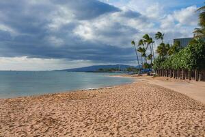 Golden sand beach with calm turquoise waters, tall palm trees, a pathway, and a distant mountain range under a partly cloudy sky in Oahu, Hawaii. photo