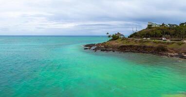 Turquoise waters meet a rocky shoreline on Oahu, Hawaii. A small hill with houses, palm trees, and a road with cars is visible under a cloudy sky. photo
