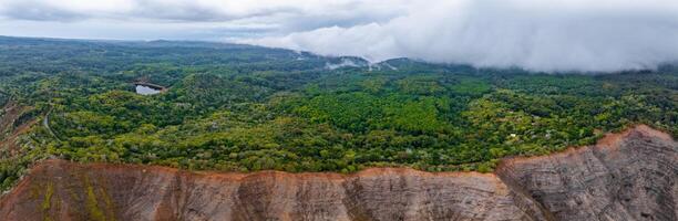 Aerial view of Kauai Island showing rugged reddish cliffs, dense green forest, a small water body, and low lying clouds on the horizon. photo