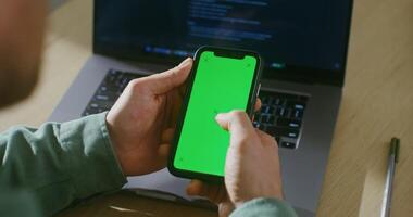 A young fair-skinned man scrolls the green screen of a mobile phone, interrupted from work on a laptop. Close-up from behind, the man's face is not visible, chroma key, copy space video