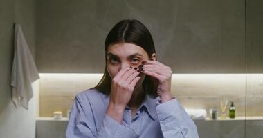 Young beautiful woman, dressed in a men's shirt, puts on moisturizing patches on the lower eyelids under the eyes, looking directly into the camera as in a mirror, standing in a stylish bathroom video