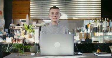 Young beautiful woman with short cropped hair, wearing glasses, with pierced ears, looking directly at the camera without smiling while sitting in front of an open laptop in a modern coffee shop video