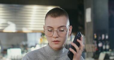 Young beautiful woman with short cut hair, wearing glasses with piercings in her ears, with a serious look answers a mobile phone call and talks drinking coffee while sitting in a coffee shop video