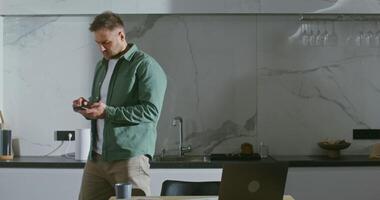 A young stylishly dressed man of European appearance with a short light beard, texting on a mobile phone, experiencing different emotions, interrupted from work on a laptop, in the kitchen at home video