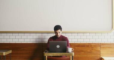 Handsome brunette man dressed in casual clothes, with wireless headphones in his ears smiling while talking on chat sitting at a table in a cozy cafe video