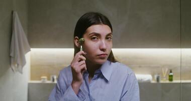 A young beautiful woman of European appearance, dressed in a man's shirt, makes a face massage with a manual roller massager, looking directly into the camera as in a mirror, in a modern bathroom video
