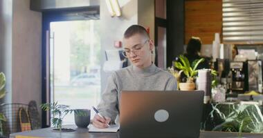 Young woman with glasses, trimmed to look like a boy, makes notes in a notebook while sitting at a table in front of an open laptop in a modern stylish coffee shop, studying online video
