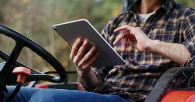 Stylishly dressed dark-haired man of European appearance uses a tablet sitting in a tractor on a farm on a sunny autumn day during a break from work, close-up, no face visible, video