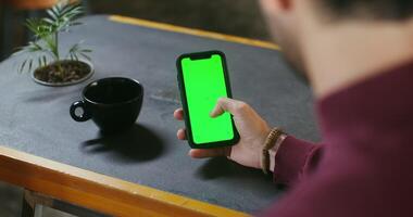 A young dark-haired man uses a green screen phone while sitting at a desk. Filming from behind, the man's face is not visible. Copy space, chroma key video