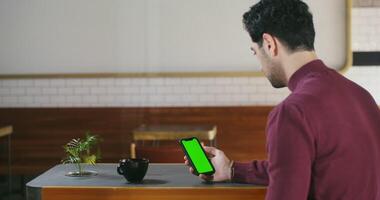 A young dark-haired man with stubble on his face uses a green screen phone while sitting at a table in a cafe. Copy space, chroma key video