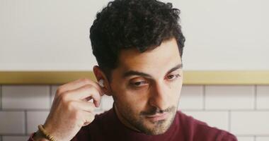 Close-up of the face of a dark-haired man plugging wireless headphones into his ears and waving his hand in greeting, smiling broadly, starting a call. video