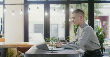 Young beautiful woman wearing glasses, dressed in casual clothes, working at a table in a modern atmospheric cafe, making notes in a notebook and using a laptop video
