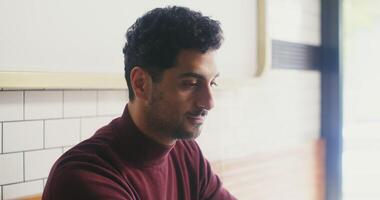 A young black-haired man with a slight unshaven face, dressed in casual clothes, brings a mug to his face and inhales the aroma of coffee with pleasure while sitting in a cafe, male face close-up video