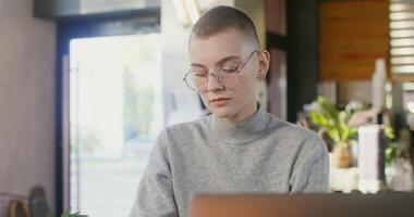 Online learning at the table in a modern coffee shop. A girl with very short hair makes notes with a pen in a notebook while sitting in front of an open laptop video